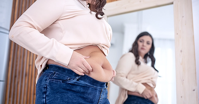 A heavyset woman looking at her tummy in the mirror | Trainest 