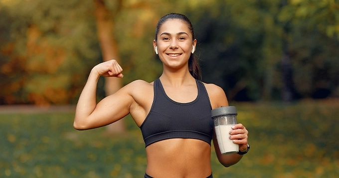 A fit woman flexing with a protein shake in hand | Trainest 