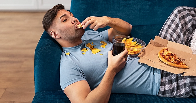 A man binge-eating junk food while laying on the couch | Trainest 