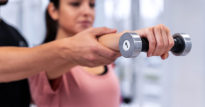 A lady lifting a light dumbbell and spotted by a trainer | Trainest 