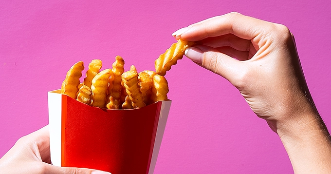 Close-up of a hand holding a box of fries and the other taking a piece | Trainest 