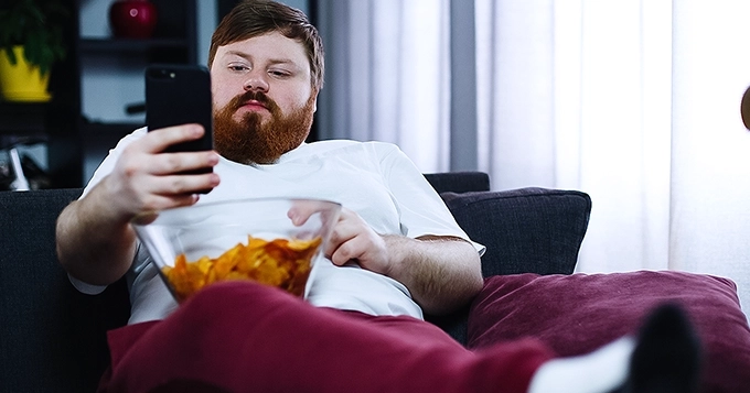 A man sitting on the couch with a bowl of chips while on his phone | Trainest 