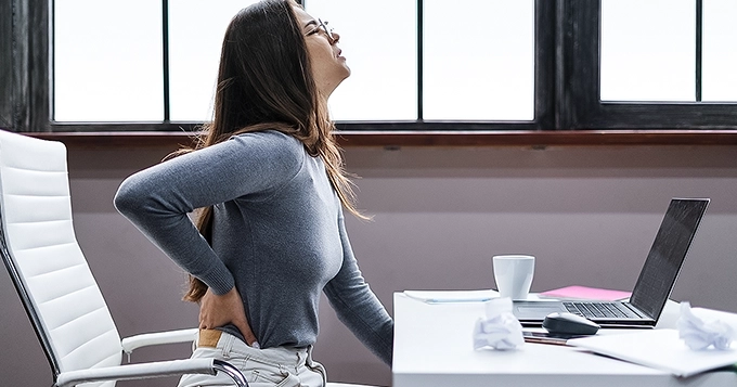 A woman in the office in pain, touching her lower back | Trainest 