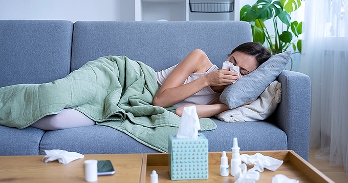 A woman lying on her couch sick and sneezing | Trainest
