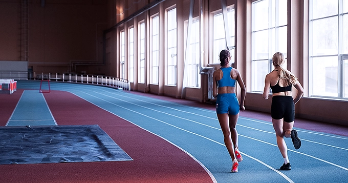 Two female athletes running around an indoor track | Trainest 