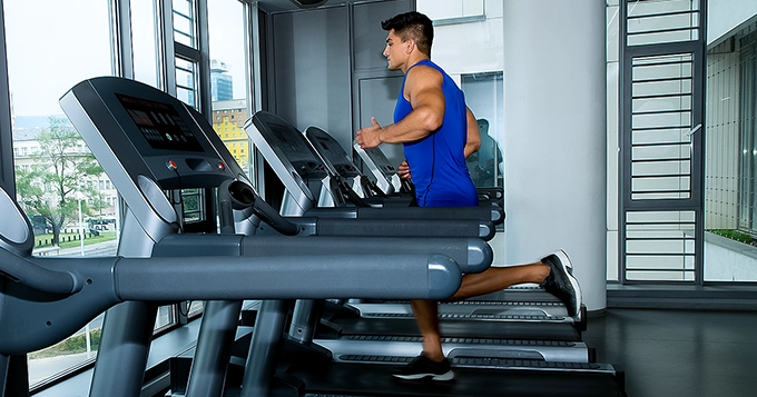 A muscular man running on a treadmill inside a commercial gym | Trainest
