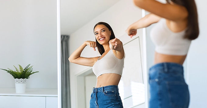 A fit woman looking at herself in the mirror, smiling | Trainest
