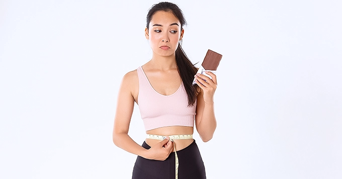 A woman holding a chocolate bar in one hand and a tape measure on her waist in another | Trainest 