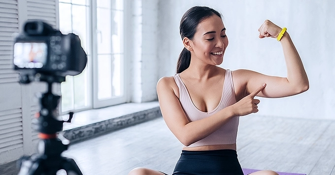 A fit woman infront of a camera pointing to her arm muscle | Trainest
