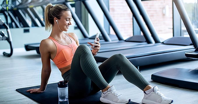 A woman at the gym looking at her phone with headphones | Trainest