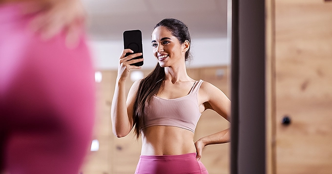 A woman taking a mirror selfie in the gym | Trainest
