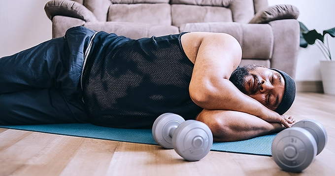 A man sleeping on the floor, surrounded by exercise equipment | Trainest
