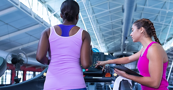A client running on a treadmill while the personal trainer is touching the control panel of the treadmill | Trainest