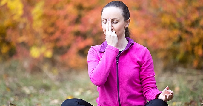 A woman meditating outdoors with one hand on her nose | Trainest
